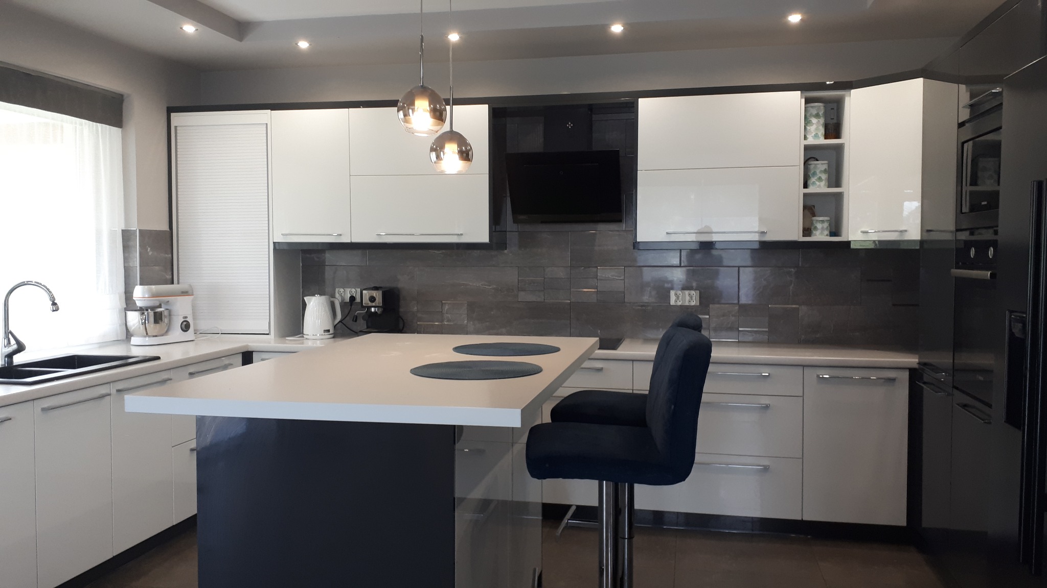 Nowoczesna kuchnia z białymi szafkami, wyspą z ciemnoniebieską podstawą i dwoma krzesłami barowymi w kolorze granatowym, oświetlona wiszącymi lampami kulami.