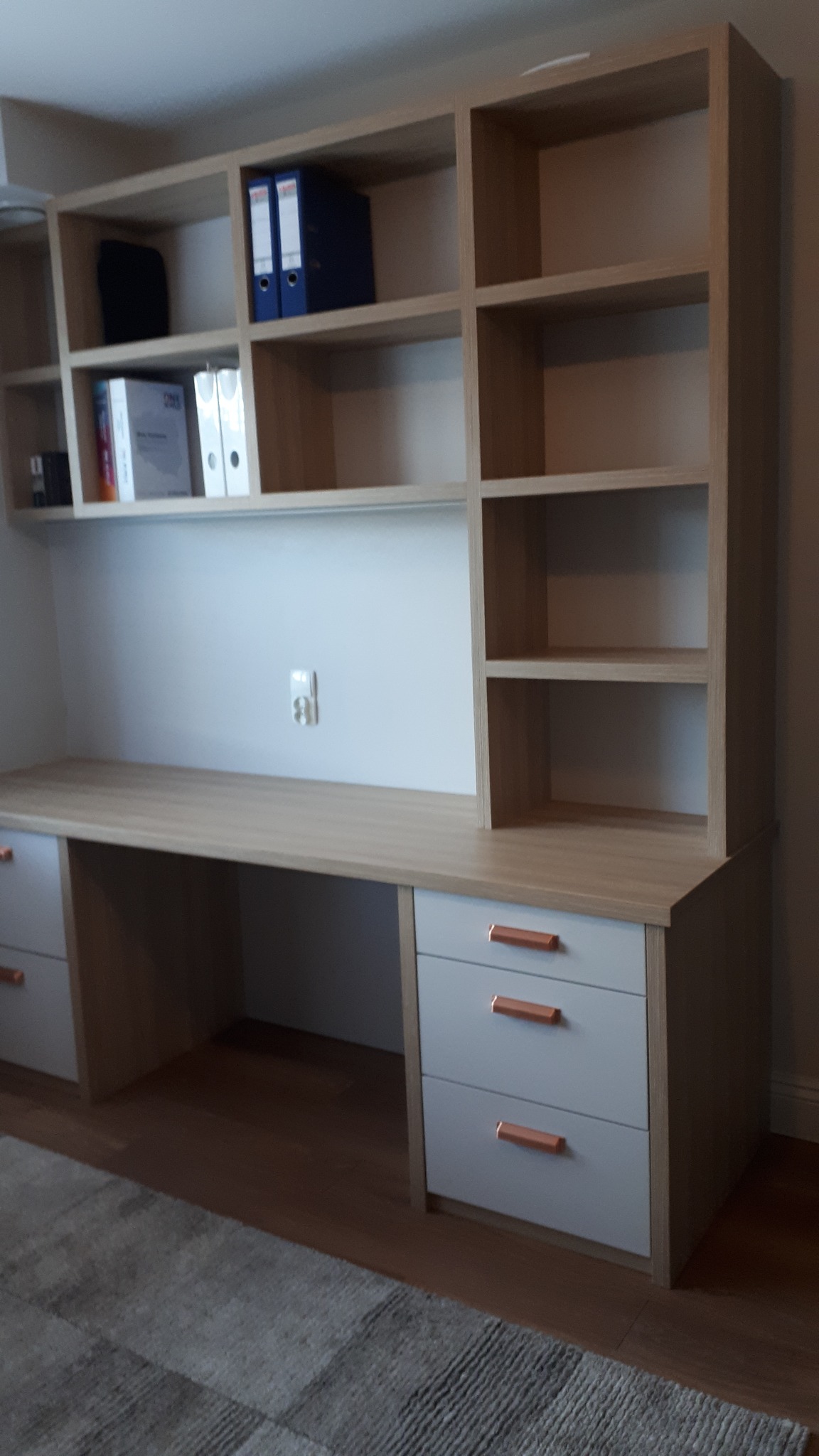 Biurko z szufladami i nadstawką z półkami, jasne drewno, szare fronty szuflad z miedzianymi uchwytami, na półkach segregatory i teczki.