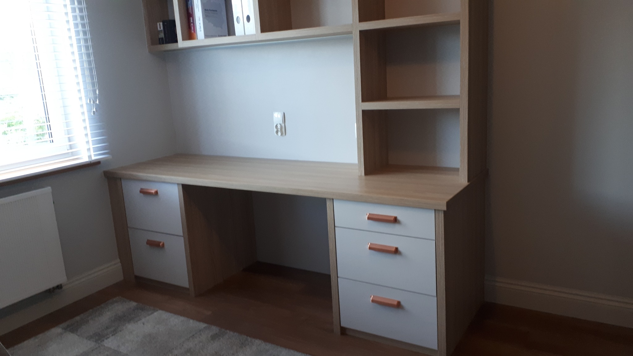 Biurko na wymiar z nadstawką z półkami, jasne drewno i białe fronty szuflad z miedzianymi uchwytami, w pokoju z roletami w oknie.