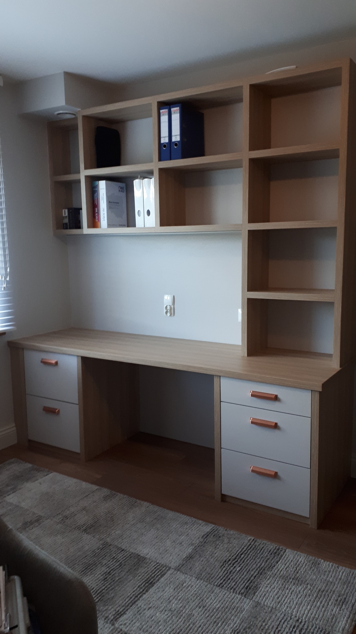 Jasny biurko z szufladami i regałem na książki, z szarymi frontami i miedzianymi uchwytami, uzupełnione o segregatory i książki, ustawione na tle jasnej ściany z gniazdkiem.