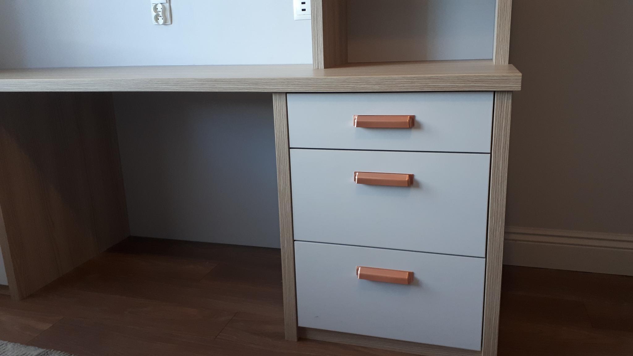 Biurko z szufladami w kolorze jasnego drewna z białymi frontami i miedzianymi uchwytami, widoczne gniazdka elektryczne na ścianie.