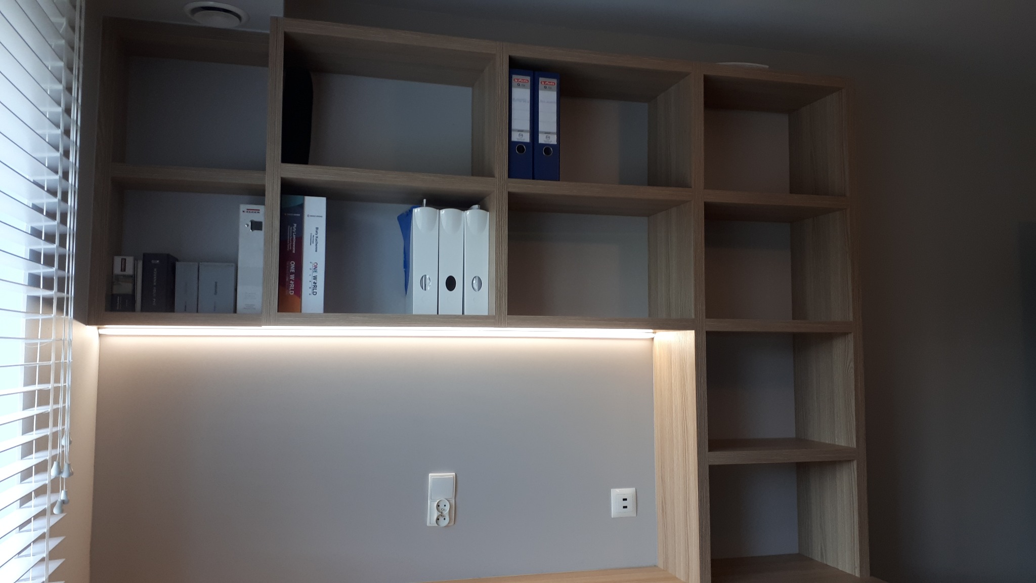 Regał drewniany z podświetleniem LED nad blatem, widoczne segregatory i książki na półkach, gniazdka elektryczne na ścianie