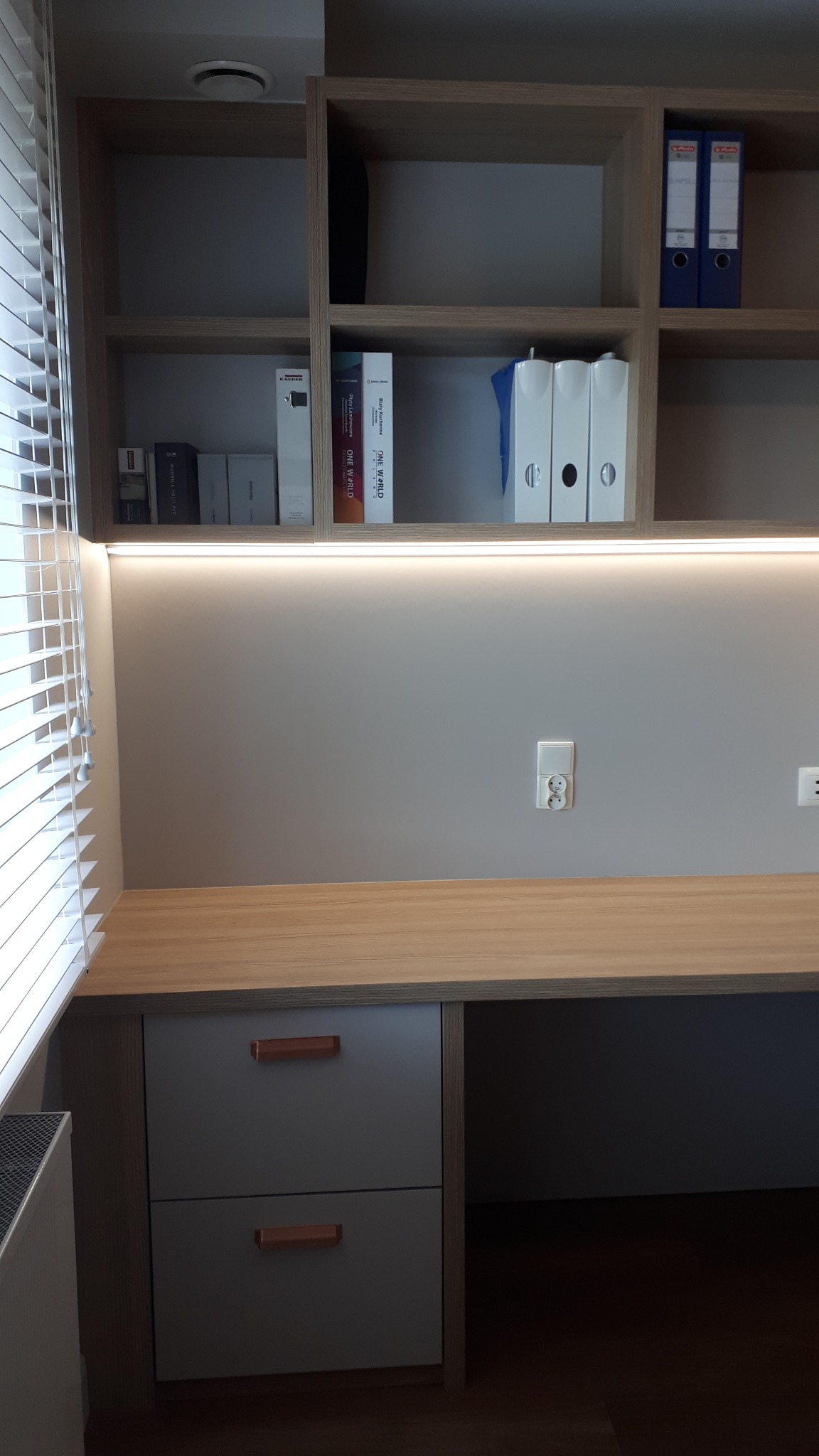 Biurko z szufladami i półkami w zabudowie, jasne drewno blatu, szare fronty szuflad z miedzianymi uchwytami, oświetlenie LED pod półkami, rolety w oknie po lewej.