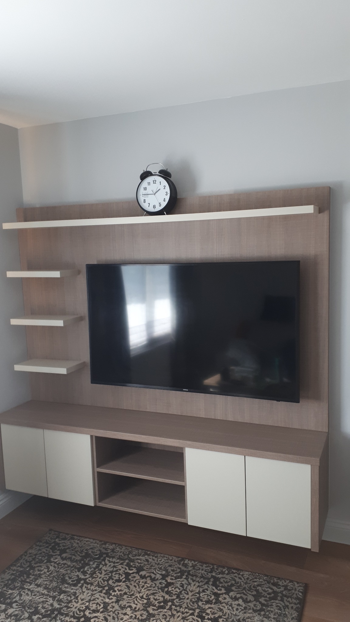 Zabudowa ściany telewizyjnej z szafkami, półkami i panelem w odcieniach beżu i bieli, z czarnym zegarem na górnej półce.