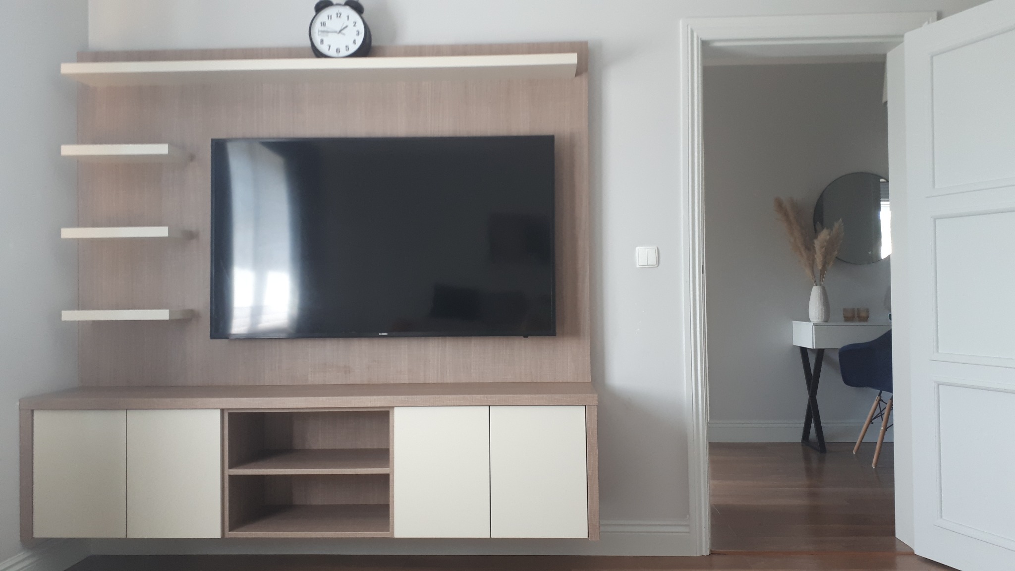 Zabudowa RTV na wymiar z szafkami i półkami w kolorze drewna i bieli, z czarnym zegarem na górnej półce, widok na wnętrze pokoju przez otwarte drzwi.