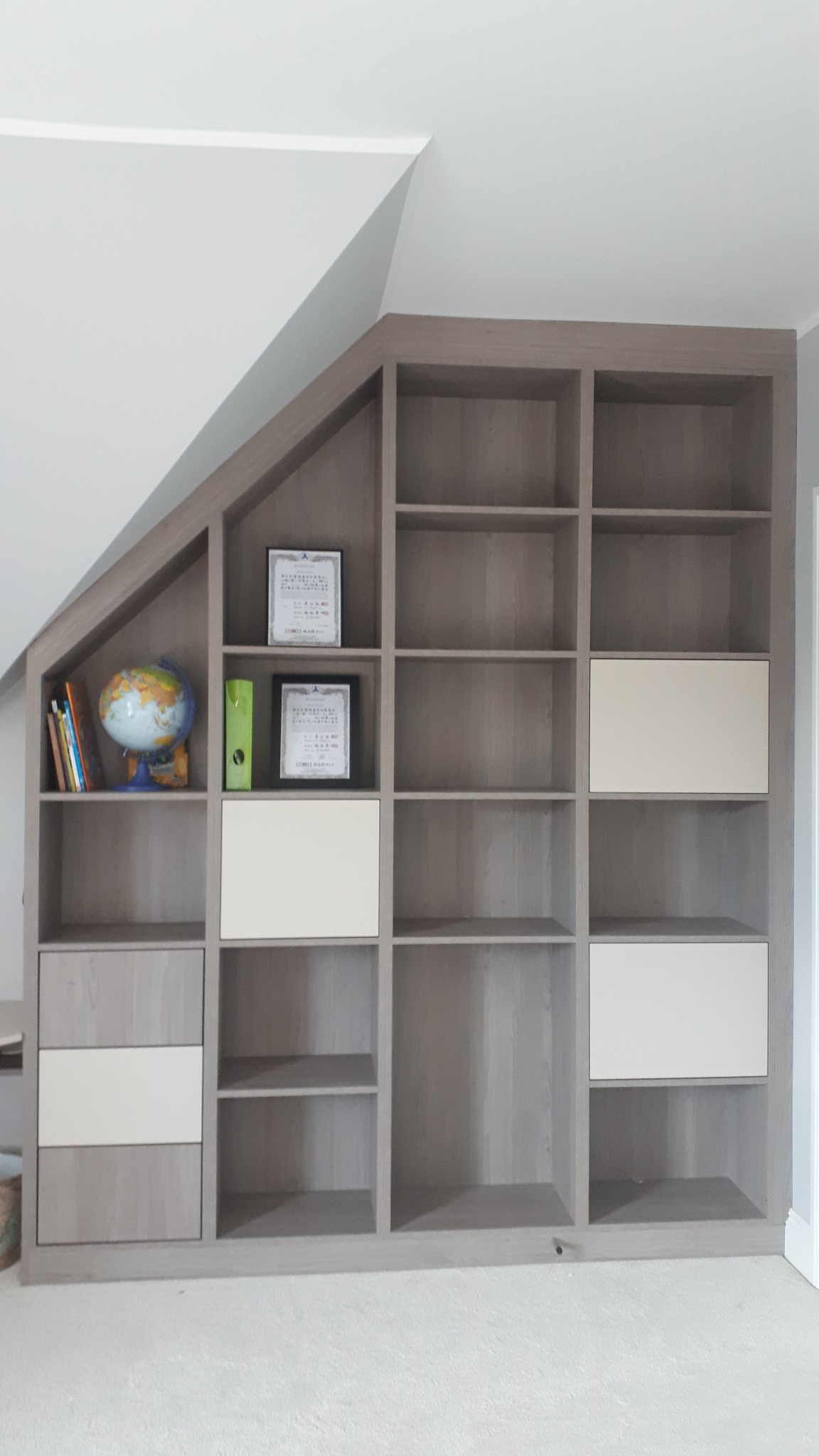 Zabudowa wnęki pod skosem z półkami i szafkami w odcieniach szarości i bieli, na półkach książki, globus, segregatory i ramki z certyfikatami.