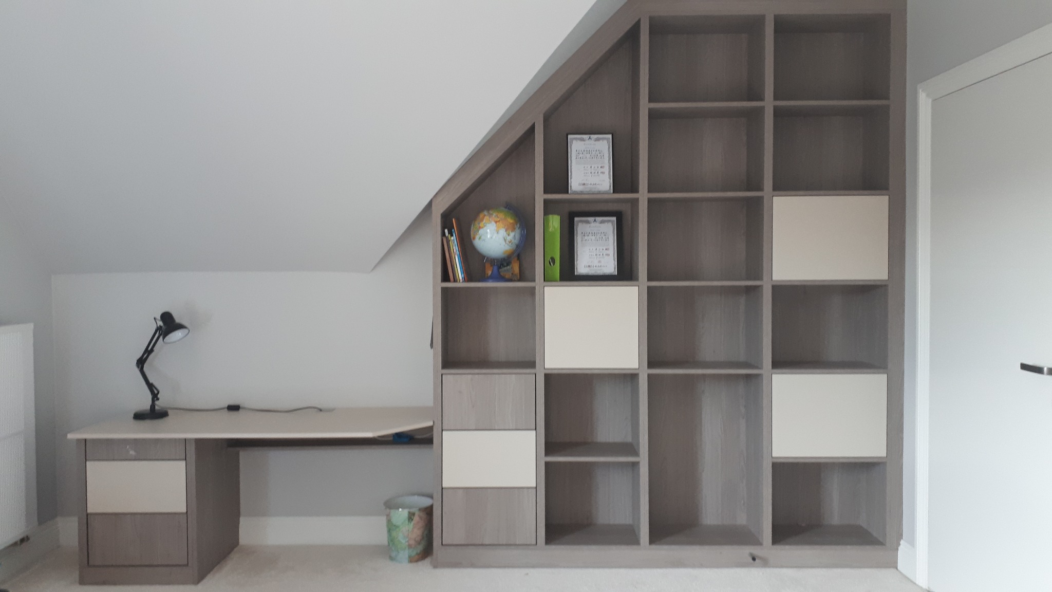Zabudowa pod skosem z regałem i biurkiem w odcieniach szarości i beżu, z globusem i dekoracyjnym koszem na śmieci.