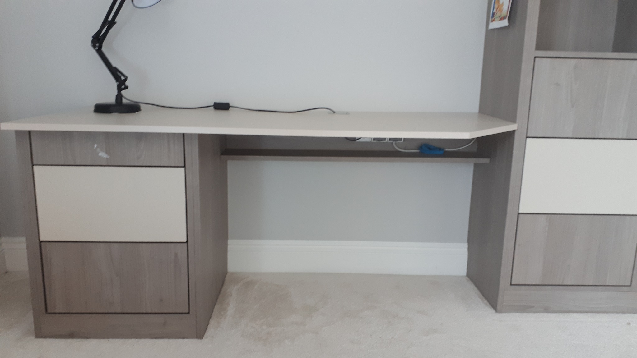 Biurko z szufladami i półką w odcieniach szarości i bieli, z lampką biurkową i widocznymi kablami, ustawione na jasnej wykładzinie.