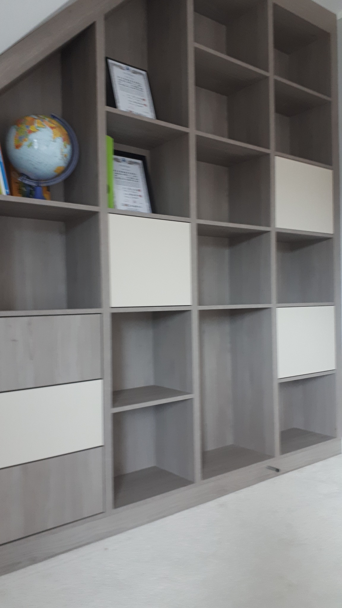 Wysoka, zabudowana szafa z półkami i szufladami w odcieniach szarości i bieli, z globusem i ramkami na zdjęciach na półkach.