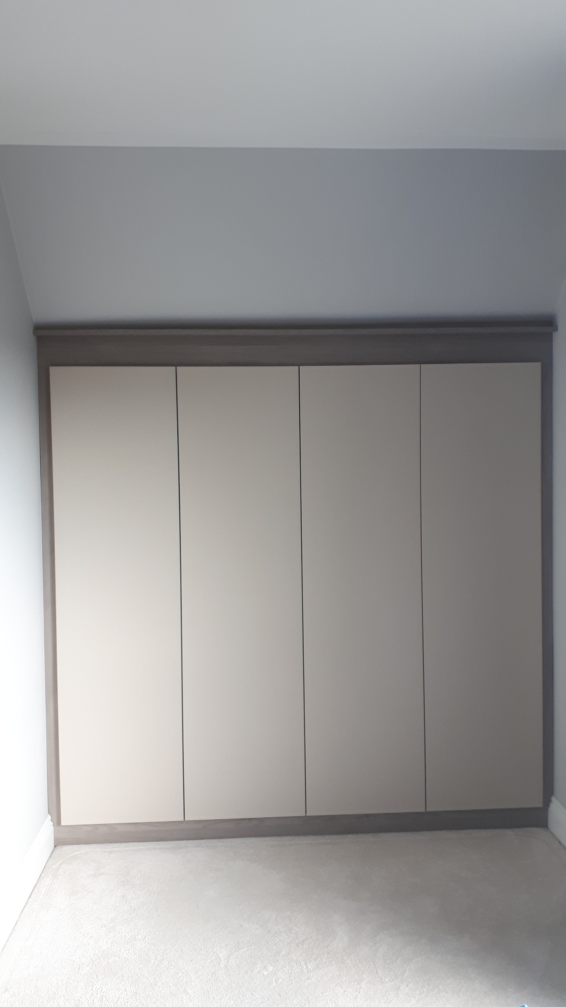 Czterodrzwiowa szafa wnękowa z jasnymi frontami i ciemniejszym obramowaniem, wkomponowana w skos pomieszczenia.