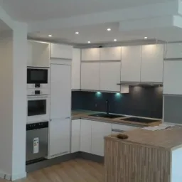 Nowoczesna, jasna kuchnia z białymi szafkami i drewnianym blatem. Oświetlenie punktowe podkreśla minimalistyczny design. Widoczne sprzęty AGD w zabudowie.