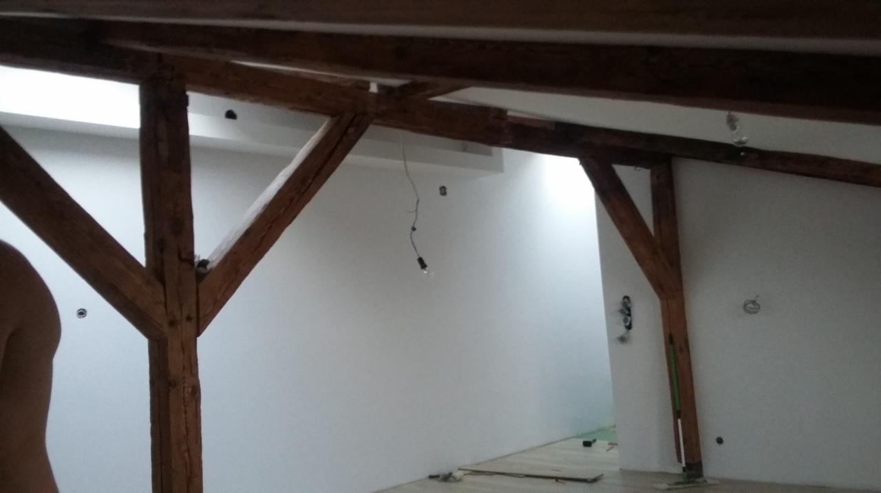 Drewniane belki stropowe w trakcie adaptacji poddasza, widoczne przewody elektryczne i świeżo pomalowane ściany na biało.