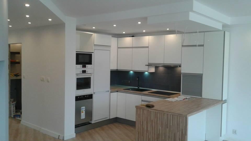 Nowoczesna, jasna kuchnia z białymi szafkami i drewnianym blatem. Oświetlenie punktowe podkreśla minimalistyczny design. Widoczne sprzęty AGD w zabudowie.