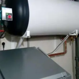 Biały bojler z czarną górną częścią, zamontowany na ścianie, z widocznymi rurami miedzianymi i szarą obudową pieca poniżej.