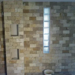 Ściana wyłożona trawertynem z wnękami i luksferami. Widoczny kubeł na podłodze. Wnętrze w trakcie wykańczania, widoczne niedoskonałości i brak fug.