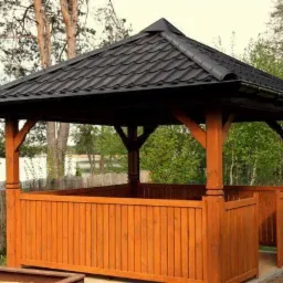 Drewniana altana ogrodowa z czarnym, spadzistym dachem z dachówki, widoczna na tle jeziora i drzew.