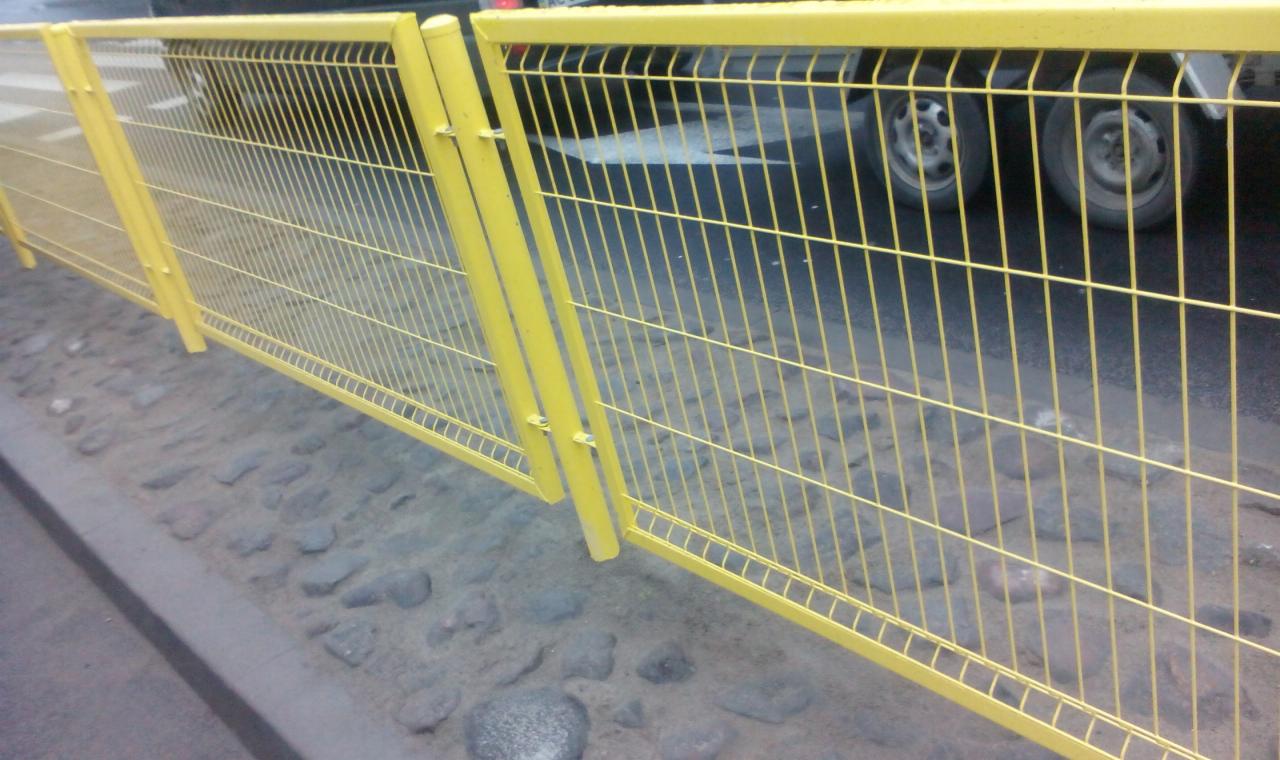 Żółte, metalowe ogrodzenie tymczasowe ustawione na krawężniku z kostki brukowej, z widocznymi kołami samochodu w tle.