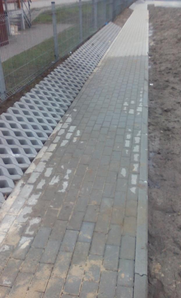 Świeżo ułożona, szara kostka brukowa sąsiaduje z ażurowymi płytami betonowymi, całość oddzielona od trawnika metalowym ogrodzeniem.