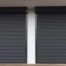 Dwie czarne rolety zewnętrzne całkowicie zasłonięte, zamontowane na elewacji budynku, widoczne poziome lamele i kasety rolet.