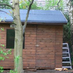 Mdach Paweł Mucha - Drewniany domek narzędziowy z ciemnym dachem, obok drzewo z budką dla ptaków i drabina. Ściany z desek w ciepłym, brązowym odcieniu, otoczone zielenią.