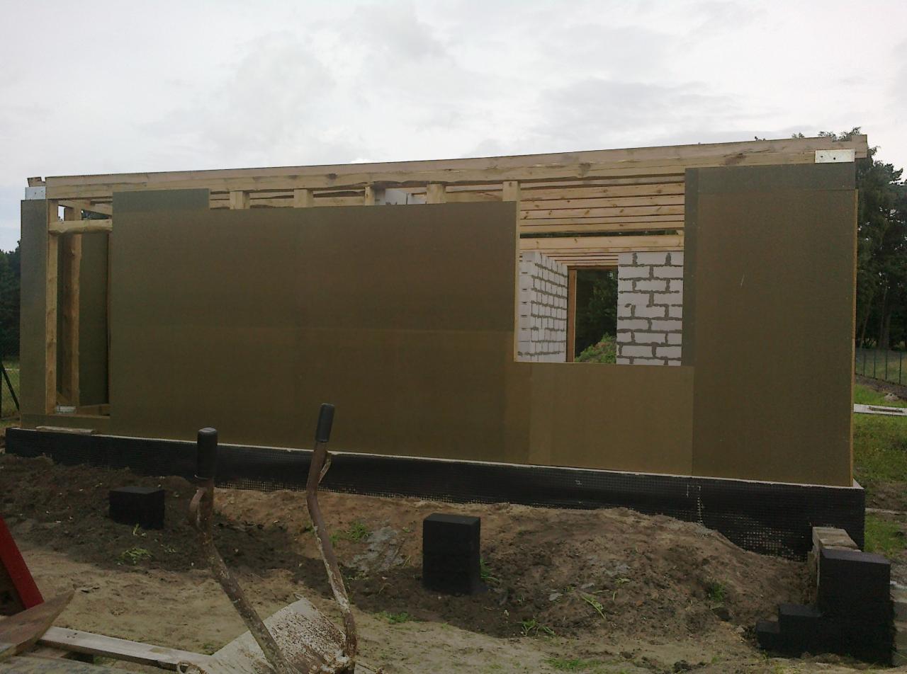 Budowa domu szkieletowego: widok ściany z izolacją termiczną, drewniana konstrukcja stropu i fragment ściany z pustaków. Widoczne narzędzia na pierwszym planie.