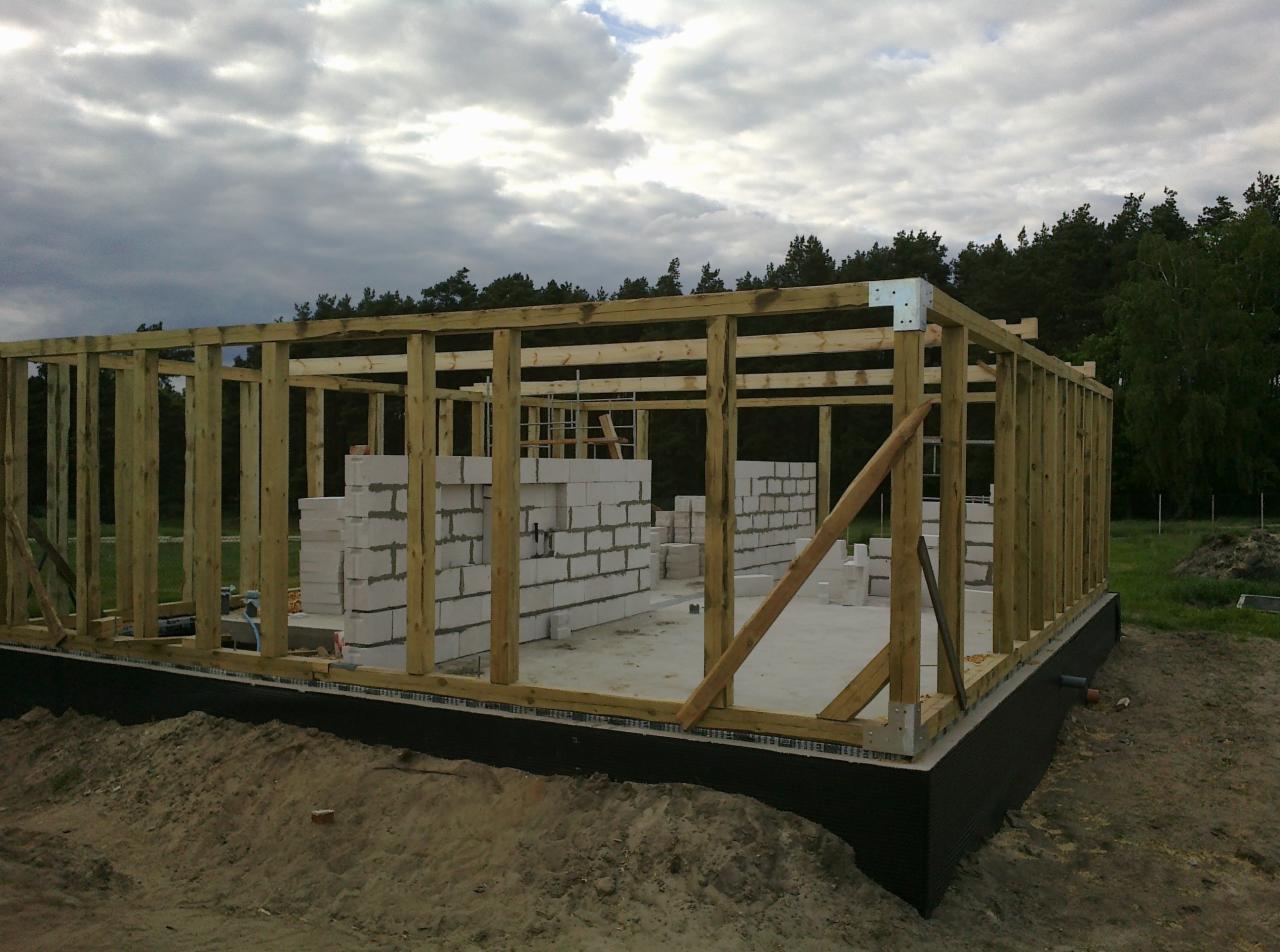 Surowy szkielet domu w budowie z drewnianą konstrukcją i ścianami z bloczków, widok zewnętrzny na tle pochmurnego nieba i drzew.