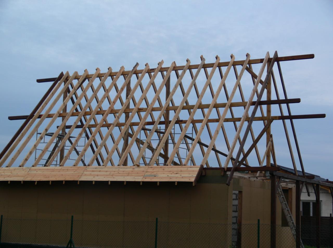 Konstrukcja drewniana dachu w trakcie budowy, widoczne krokwie i łaty, budynek w stanie surowym otwartym, ogrodzenie siatkowe w tle.
