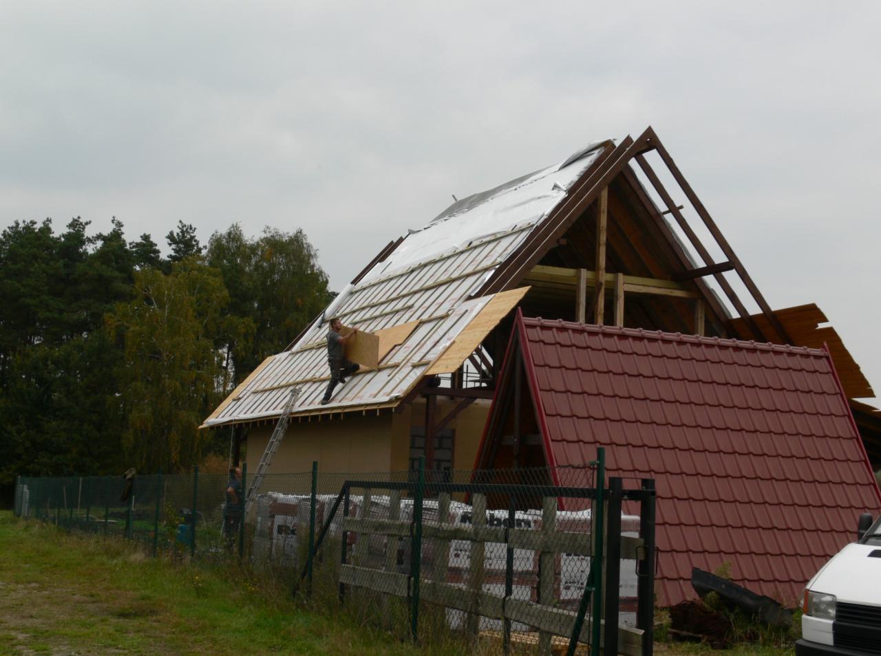 Prace dekarskie na dachu domu: montaż płyt OSB na łatach pokrytych folią. W tle widać więźbę dachową i fragment dachu z czerwoną dachówką. Pracownik na drabinie.