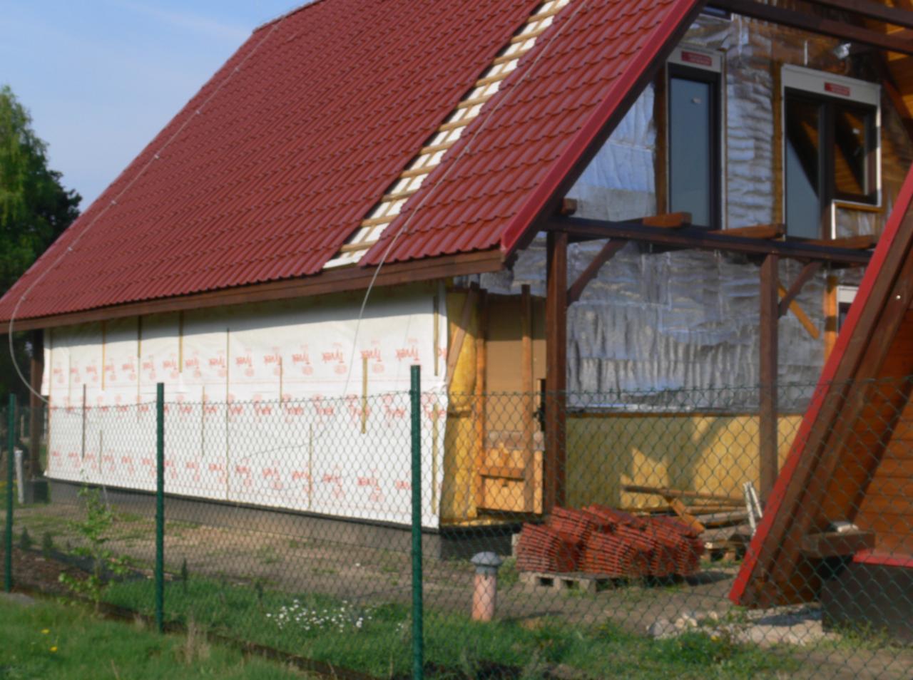 Dom w trakcie budowy, widoczna czerwona dachówka, ocieplenie ścian folią i wełną mineralną. Częściowo zadaszony taras z drewnianą konstrukcją.