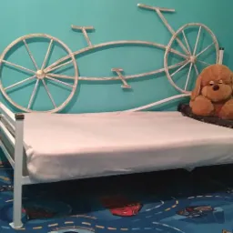 Metalowe łóżko dziecięce z oryginalnym zagłówkiem w kształcie roweru, pluszowy miś na łóżku, kolorowy dywan z motywem ulicy, turkusowa ściana w tle.