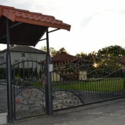 Czarna, kuta brama wjazdowa z dekoracyjnymi elementami roślinnymi, osłonięta zadaszeniem z dachówką. Widok na posesję i budynki w tle, oświetlone popołudniowym słońcem.