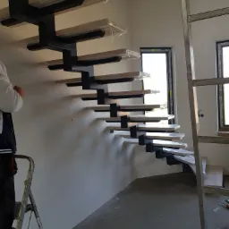 Wnętrze w trakcie wykańczania z zamontowanymi schodami dywanowymi z drewnianymi stopniami zabezpieczonymi folią i czarną metalową konstrukcją nośną, widoczne okna i drabina.