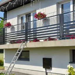 Balkon z nowoczesną, poziomą balustradą w kolorze antracytowym, ozdobiony wiszącymi donicami z różowymi kwiatami, drabina oparta o balkon.