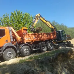 Pomarańczowa ciężarówka i koparka Liebherr na nierównym terenie, załadunek ziemi w słoneczny dzień. Widoczny fragment wykopu i zieleń w tle.