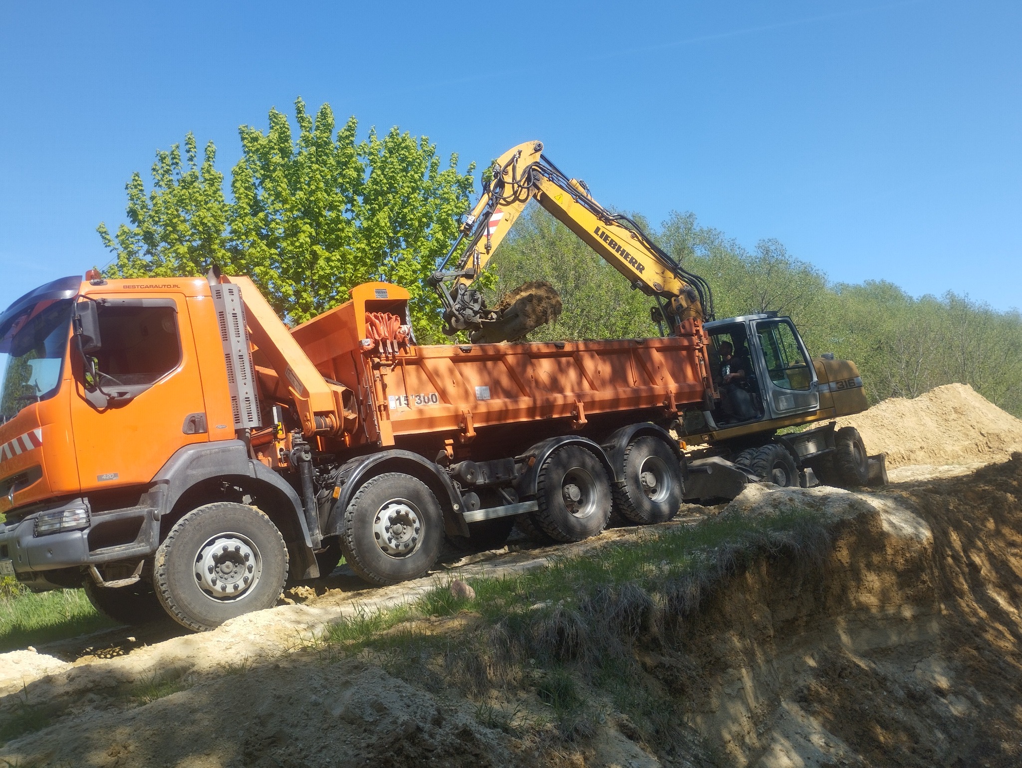Pomarańczowa ciężarówka i koparka Liebherr na nierównym terenie, załadunek ziemi w słoneczny dzień. Widoczny fragment wykopu i zieleń w tle.