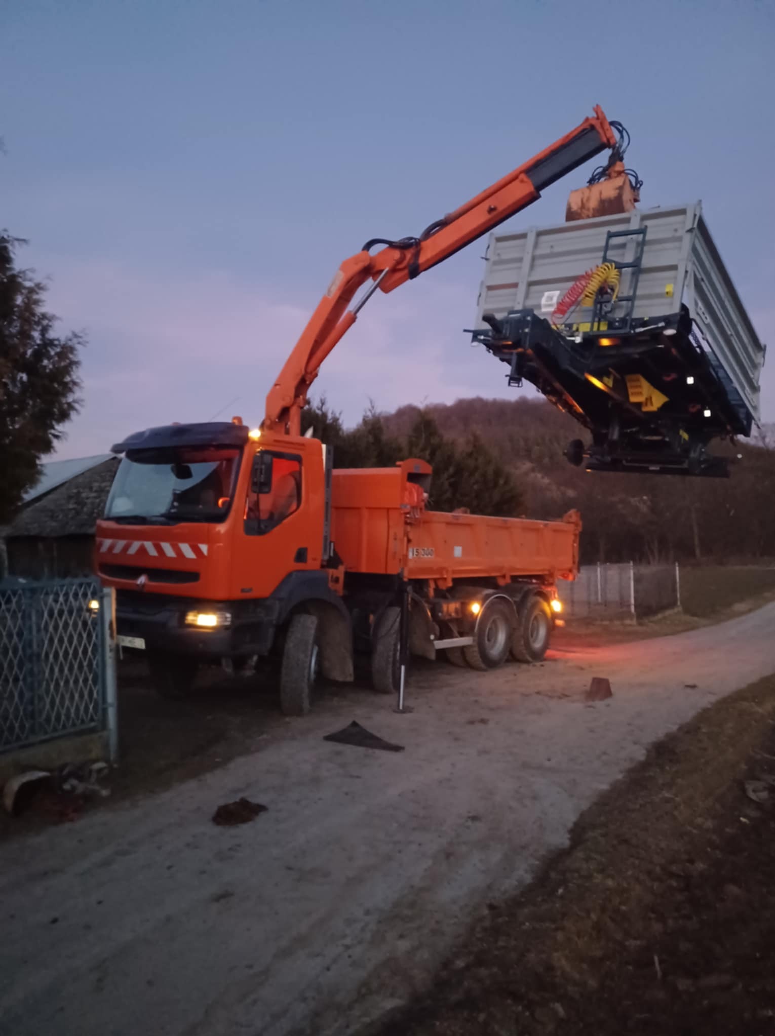 Pomarańczowa ciężarówka z dźwigiem podnosi przyczepę na tle drzew i nieba o zmierzchu. Widoczne oświetlenie i elementy konstrukcyjne pojazdów.