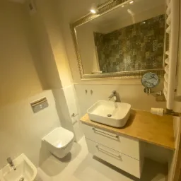 Nowoczesna łazienka z białą armaturą, kwadratową umywalką na drewnianym blacie, wiszącą toaletą i bidetem, oraz lustrem w złotej ramie odbijającym ścianę z kolorowymi płytkami.