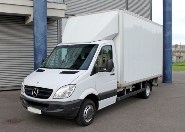 Biały samochód dostawczy Mercedes-Benz Sprinter z zabudową kontenerową na tle szarej fasady budynku i niebieskiego słupa.