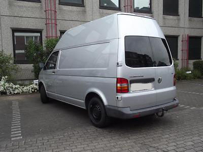 Srebrny bus z wysokim dachem zaparkowany na brukowanym parkingu przed budynkiem z szarych płyt. Widok z tyłu, zarys roślinności po lewej stronie.