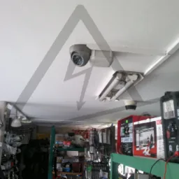 Kamera monitoringu kopułkowa zamontowana na suficie obok lampy fluorescencyjnej. W tle widoczne regały z narzędziami i akcesoriami.