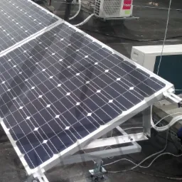 Panele fotowoltaiczne na dachu budynku, zamontowane na metalowej konstrukcji, z widoczną jednostką zewnętrzną klimatyzacji. Instalacja w trakcie prac, widoczna odzież robocza.