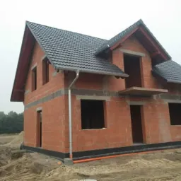 Dwukondygnacyjny dom w stanie surowym otwartym z czarną dach&oacute;wką, widoczne pustaki ceramiczne i przygotowane otwory okienne. Na pierwszym planie piasek.