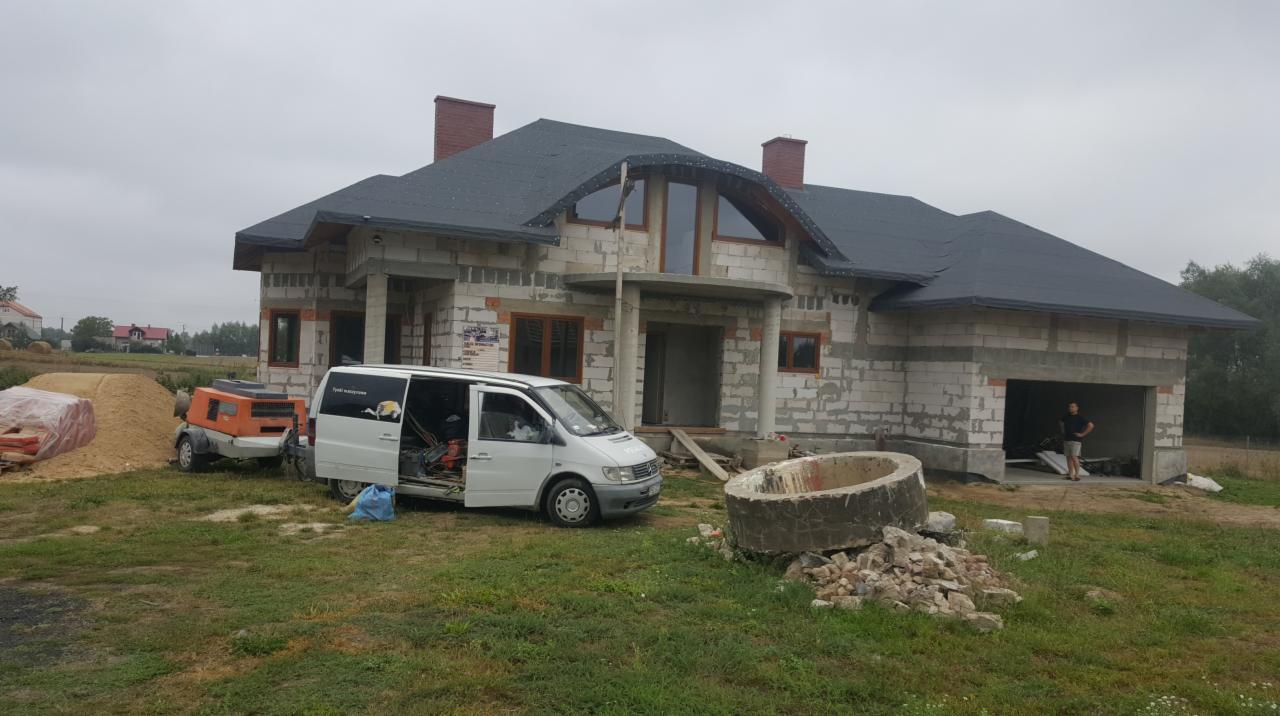 Niedokończony dom z pustaków, z widocznym sprzętem budowlanym, otwartym busem i stertą gruzu na trawniku.