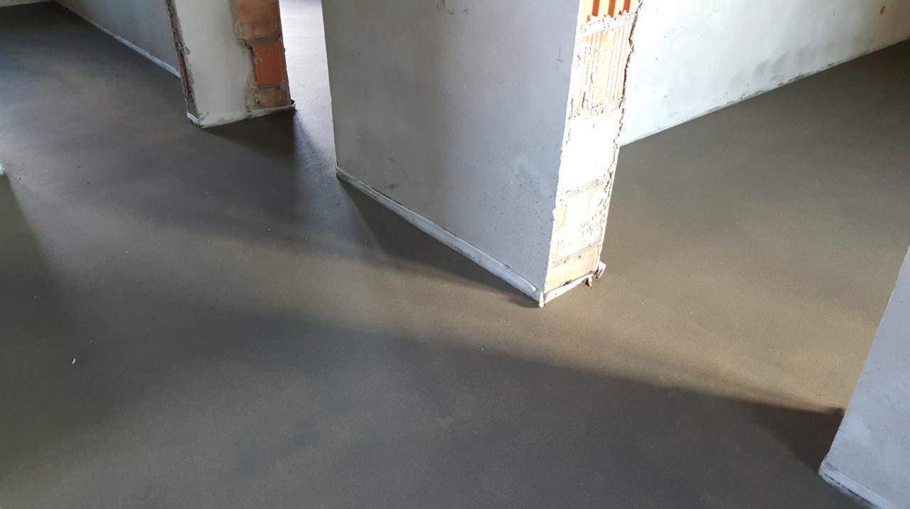 Świeżo wylana, gładka, szara podłoga w surowym wnętrzu, z widocznymi fragmentami ceglanych ścian i niedokończonymi ścianami działowymi.