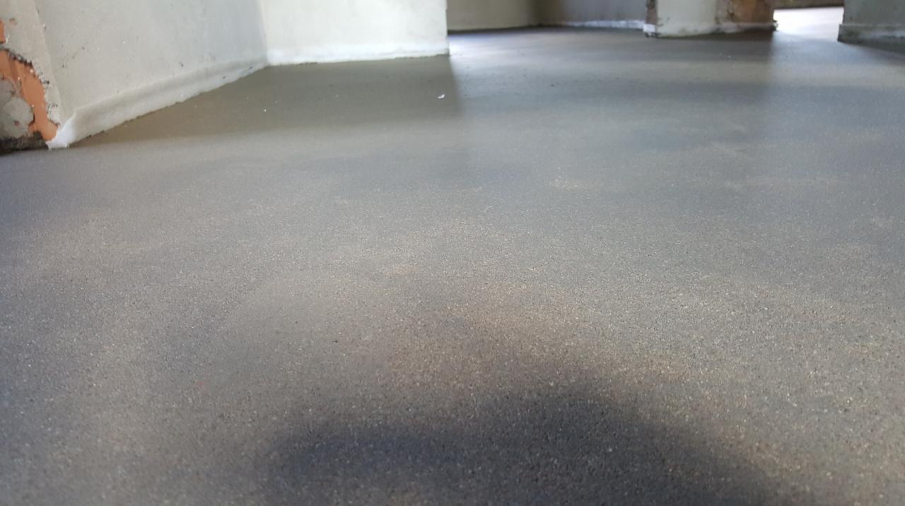 Świeżo wylana, gładka, szara podłoga z widocznymi śladami wilgoci, przy ścianie zabezpieczona białą pianką dylatacyjną, z surowymi fragmentami muru w rogu.