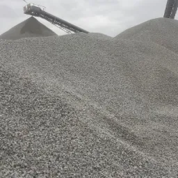 Góra szarego grysu 16-22 pod taśmociągiem na tle pochmurnego nieba. Widok z dołu, ukazujący fakturę kamieni i konstrukcję taśmociągu.