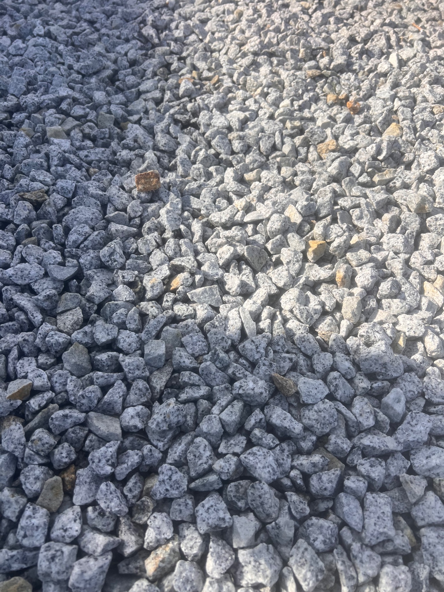 Szare, drobne kamienie z granitowym wzorem, oświetlone z prawej strony, tworzące teksturę powierzchni. Pojedyncze, brązowe kamienie urozmaicają tło.