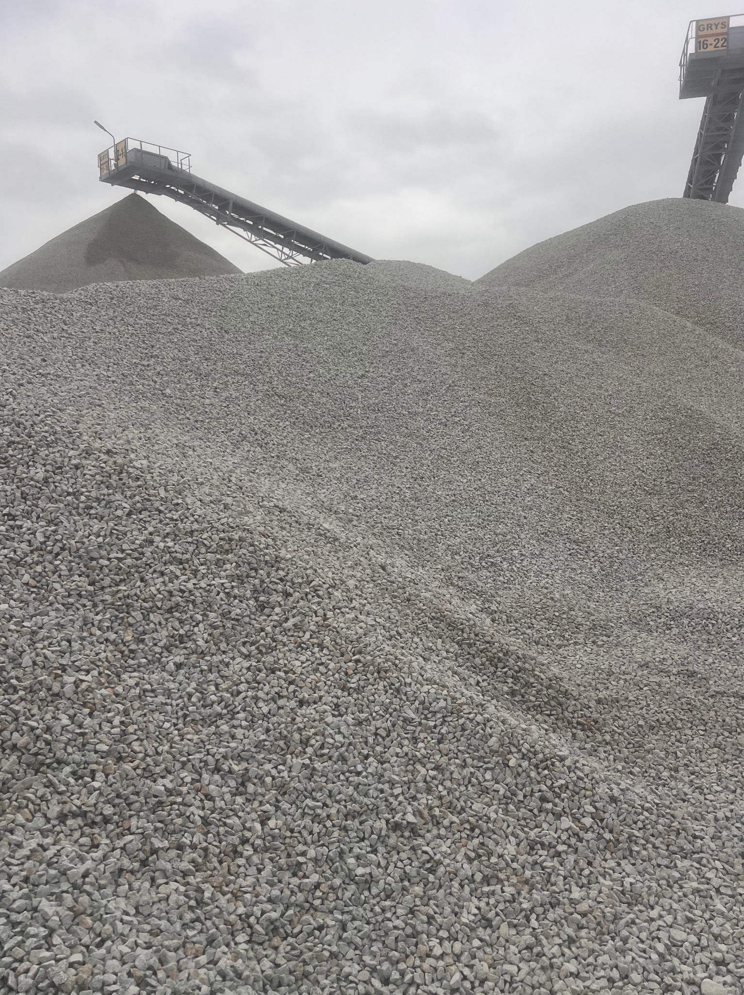 Góra szarego grysu 16-22 pod taśmociągiem na tle pochmurnego nieba. Widok z dołu, ukazujący fakturę kamieni i konstrukcję taśmociągu.