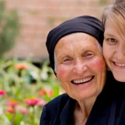 Uśmiechnięta starsza kobieta w czarnej chuście, obejmowana przez młodą dziewczynę na tle kwitnącego ogrodu z r&oacute;żowymi i pomarańczowymi kwiatami.