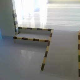 Wykończone schody z rampą w jasnym wnętrzu, zabezpieczone listwami w brązowo-beżową szachownicę, na błyszczącej podłodze. Detal wykończenia.